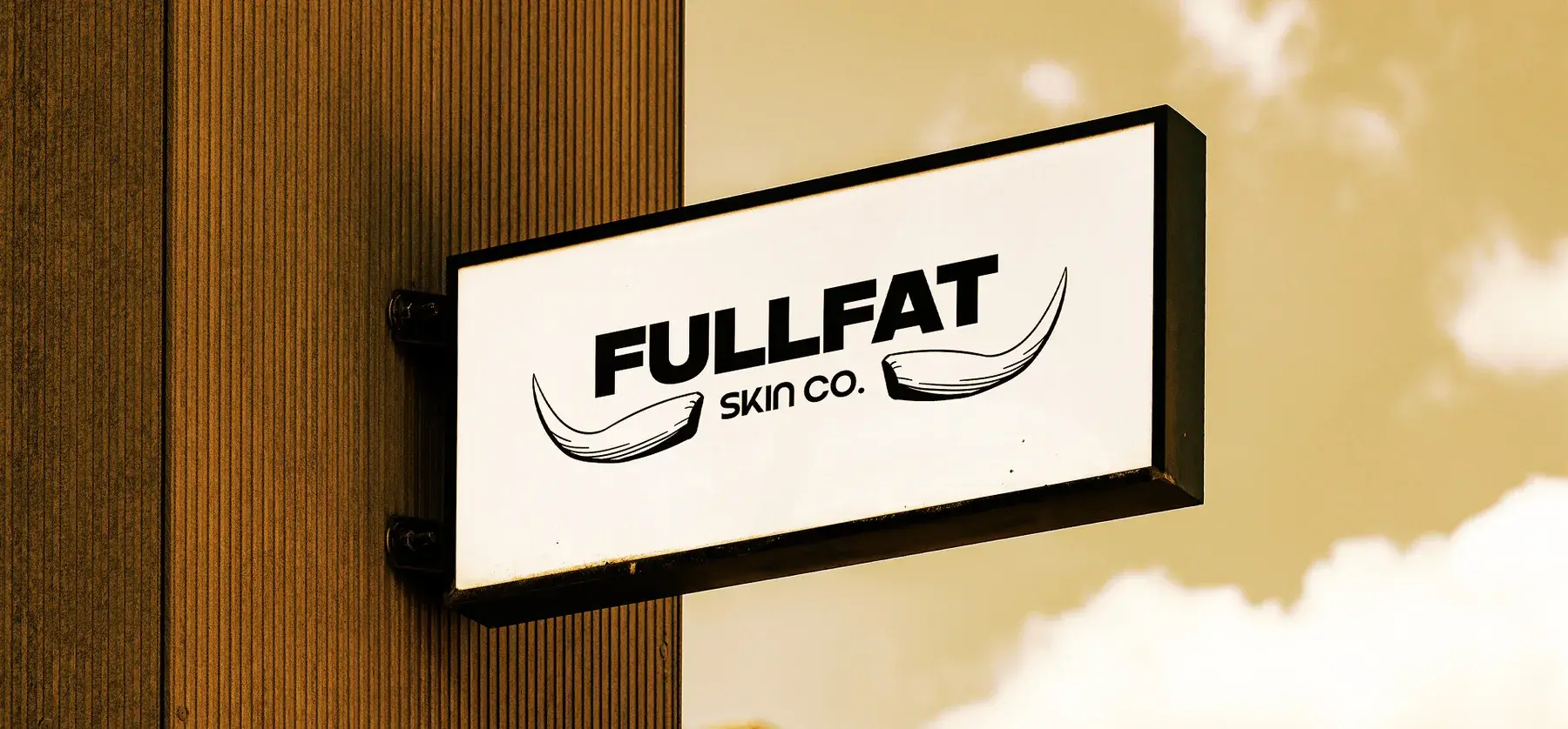 A wall-mounted lightbox sign with the black FULLFAT Skin Co. logo against a warm sky